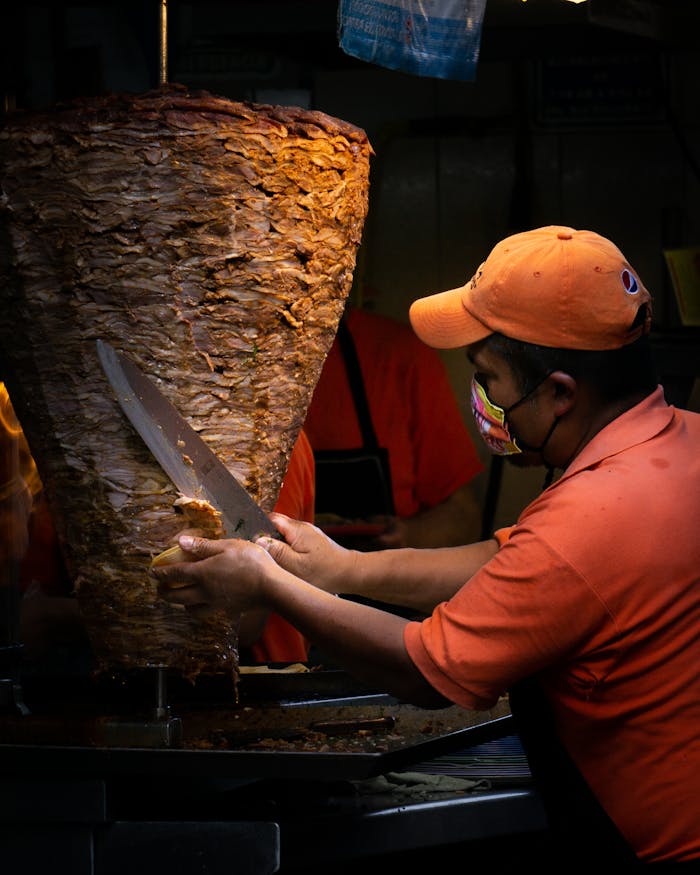 A man slices seasoned al pastor meat from a vertical spit in a busy food stall.